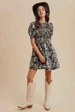 Load image into Gallery viewer, Woman wearing a floral dress with puffed sleeves, white cowboy boots, and a black hat on a beige background