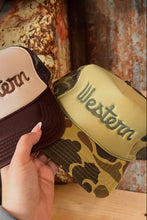 Load image into Gallery viewer, Two 'Western' branded hats, one brown and one with a camouflage pattern, held by a hand against a rustic background.