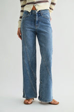 Load image into Gallery viewer, A person wearing high-waisted, wide-leg blue denim jeans with diagonal seam details down the front of the legs, styled with a cream striped long-sleeve top and brown sandals against a light gray studio background.