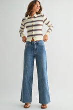 Load image into Gallery viewer, A woman modeling a cream knit button-front cardigan with dark brown and black horizontal stripes, paired with wide-leg blue jeans and brown sandals, against a light gray studio background.