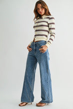 Load image into Gallery viewer, A woman modeling a cream ribbed knit long-sleeve polo shirt with dark brown and black horizontal stripes, paired with high-waisted, wide-leg blue denim jeans and brown heeled sandals, against a light gray studio background.
