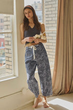 Load image into Gallery viewer, Woman wearing a dark gray sleeveless top and patterned pants standing by a window.