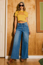 Load image into Gallery viewer, Woman wearing a yellow crop top and blue jeans standing against a wooden wall.