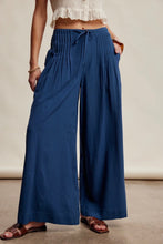 Load image into Gallery viewer, A person wearing wide-leg pleated pants in a medium-to-dark blue tone, paired with a white ruffled crop top and brown sandals, standing against a plain beige studio wall.
