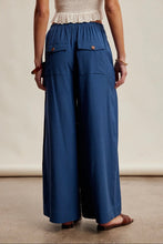 Load image into Gallery viewer, A person shown from the back wearing wide-leg blue pants with an elastic waistband, flap pockets, and brown flat sandals against a plain beige studio background.