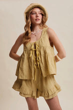 Load image into Gallery viewer, A woman modeling a matching two-piece linen romper in a warm mustard yellow color, featuring a tie-front top and ruffled shorts, styled with a straw hat against a solid beige background.