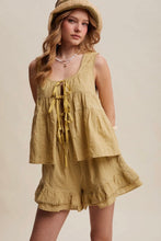 Load image into Gallery viewer, A woman wearing a matching two-piece linen romper in a warm mustard yellow color, featuring a tie-front top and ruffled shorts, styled with a straw hat against a solid beige background.