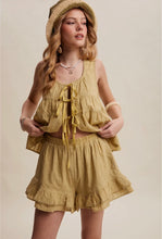 Load image into Gallery viewer, A woman modeling a matching two-piece set consisting of a sleeveless tie-front ruffled top and matching ruffled shorts in a warm mustard yellow color, styled with a woven straw hat and layered necklaces against a solid beige background.