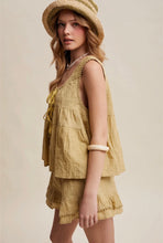 Load image into Gallery viewer, A woman wearing a light beige linen-look two-piece set consisting of a sleeveless tiered top with tie details and matching ruffled shorts, styled with a straw hat against a solid beige background.