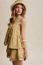 Load image into Gallery viewer, A woman wearing a matching yellow linen tiered romper with ruffle trim, a straw hat, and a pearl necklace, photographed against a solid beige studio background.