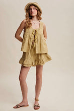 Load image into Gallery viewer, A woman modeling a light beige linen two-piece set consisting of a sleeveless tiered top with a front tie and matching ruffled shorts, styled with a straw hat and sandals against a solid beige background.