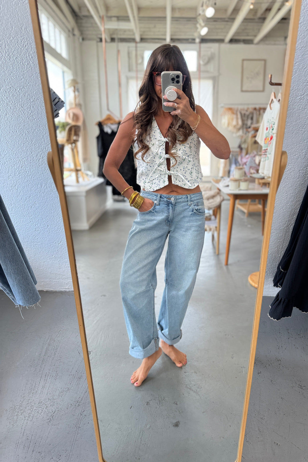 Person taking a mirror selfie wearing a white patterned top and light blue jeans in a room.