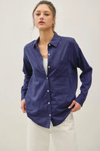 Load image into Gallery viewer, A woman modeling a navy blue long-sleeve button-up shirt with a chest pocket, worn open over a white top and paired with white pants, against a plain light gray studio background.