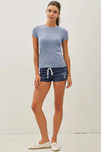 Load image into Gallery viewer, A woman modeling a light blue short-sleeve crewneck t-shirt and navy blue athletic shorts with a white drawstring, paired with white slip-on shoes, against a plain off-white studio background.