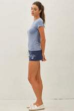 Load image into Gallery viewer, A woman standing in profile wearing a light blue short-sleeve t-shirt, navy blue athletic shorts with a white graphic print, and white slip-on shoes against a plain white studio background.
