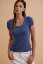 Load image into Gallery viewer, A woman wearing a fitted short-sleeve scoop-neck t-shirt in a dusty medium blue, paired with white pants, standing against a solid beige studio background.