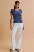 Load image into Gallery viewer, A woman modeling a short-sleeve scoop-neck knit top in dusty blue, paired with white wide-leg pants and gray sandals, against a solid beige studio background.