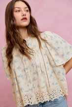 Load image into Gallery viewer, A woman wearing a cream-colored short-sleeve blouse with a light blue floral print, eyelet lace trim, and a front zipper, paired with blue jeans against a solid pink background.