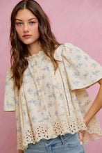 Load image into Gallery viewer, A woman wearing a cream-colored short-sleeve blouse with a light blue floral print and eyelet lace trim, paired with light-wash blue jeans, set against a solid pink background.