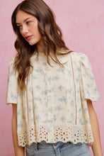 Load image into Gallery viewer, A woman wearing a short-sleeve cream eyelet blouse with a light blue floral print, paired with light-wash jeans, set against a solid pink background.
