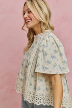 Load image into Gallery viewer, A woman wearing a short-sleeve cream-colored blouse with a light blue floral print, eyelet lace trim at the collar and hem, and a lace-up front, posed against a solid pink background.