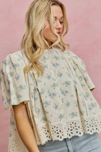 Load image into Gallery viewer, A woman wearing a cream-colored short-sleeve blouse with a light blue floral print, eyelet lace trim, and ruffle details, paired with light-wash jeans against a solid pink background.