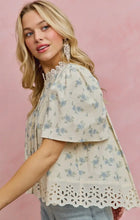 Load image into Gallery viewer, A woman wearing a short-sleeve cream blouse with a light blue floral print and eyelet lace trim, paired with light-wash jeans against a soft pink background.