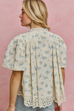 Load image into Gallery viewer, A woman shown from the back wearing a cream-colored blouse with a light blue floral print, pleated back, and eyelet lace trim, paired with light-wash jeans against a solid pink background.