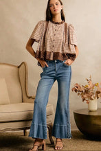 Load image into Gallery viewer, A woman models a brown and beige gingham short-sleeve blouse with a ruffled neckline and lace hem, paired with blue flare jeans with lace side detailing, in a styled indoor setting.