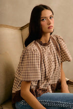 Load image into Gallery viewer, A woman wearing a brown and beige gingham blouse with lace trim and puff sleeves, paired with blue jeans, sitting on a beige armchair.