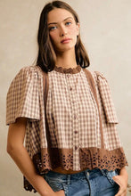 Load image into Gallery viewer, A woman wearing a brown and beige gingham short-sleeve blouse with lace trim and button-front detailing, paired with blue jeans, against a neutral beige background.