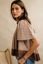 Load image into Gallery viewer, A woman wearing a brown and beige plaid short-sleeve blouse with lace trim and ruffled sleeves, paired with blue jeans, photographed against a neutral beige wall.