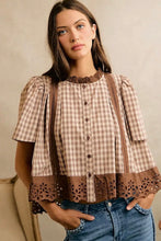 Load image into Gallery viewer, A woman wearing a brown and beige gingham short-sleeve blouse with brown lace trim and button-front detailing, paired with blue jeans, against a neutral beige background.