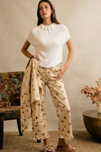 Load image into Gallery viewer, A woman stands indoors wearing a white short-sleeve knit top and beige patterned pants, holding a matching white cardigan draped over one arm. The room has a textured beige wall, a patterned chair, and a round table with dried flowers.