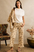 Load image into Gallery viewer, A woman models a beige patterned pants set featuring a white ruffled mock neck top and a matching beige patterned cardigan draped over her shoulder, styled in a warm, neutral-toned room.
