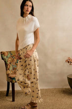 Load image into Gallery viewer, A woman stands indoors wearing a cream short-sleeve knit top and beige wide-leg pants with a small bird print. She is posed in a neutral room with a textured wall, a patterned chair, and a small table with flowers.