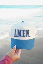 Load image into Gallery viewer, A hand holds a two-tone trucker hat with 'AMEN.' embroidered on the front, set against a blurred beach background with ocean waves.