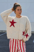 Load image into Gallery viewer, A woman wearing a fuzzy cream cardigan with red star motifs and red-and-white striped pants, standing outdoors against a light wall.