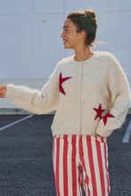Load image into Gallery viewer, A woman wearing a fuzzy cream cardigan with red star motifs and red-and-white striped pants, standing outdoors against a light gray wall.