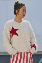 Load image into Gallery viewer, A woman wearing a fuzzy cream cardigan with red star details and red-and-white striped pants, standing against a pale wall.