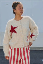 Load image into Gallery viewer, A woman wearing a fuzzy cream cardigan with red star patches and red-and-white striped pants, standing outdoors against a light wall.