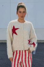 Load image into Gallery viewer, A woman wearing a fuzzy cream cardigan with red star patterns and red-and-white striped pants, standing outdoors against a light gray wall.