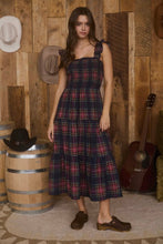 Load image into Gallery viewer, Woman in a plaid dress standing in front of a wooden wall with hats and a guitar.