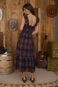 Woman in a plaid dress standing in a rustic setting with wooden wall, guitar, and hay.