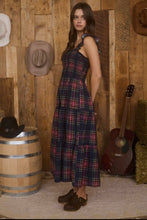Load image into Gallery viewer, Woman in a plaid dress standing against a wooden wall with hats and a guitar.