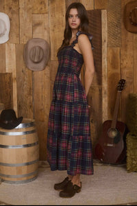 Woman in a plaid dress standing against a wooden wall with hats and a guitar.