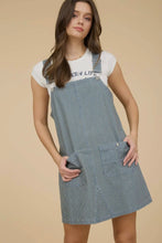 Load image into Gallery viewer, Woman wearing a blue checkered pinafore dress over a white t-shirt with text on a beige background
