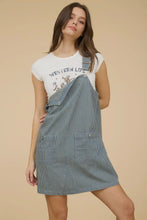 Load image into Gallery viewer, Woman wearing a white t-shirt with text and graphics, paired with a blue denim pinafore dress on a beige background.