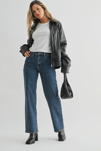 Woman wearing a black leather jacket, white shirt, blue jeans, and black boots on a light gray background