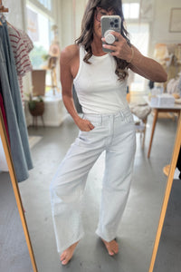 Person wearing a white tank top and light blue jeans standing in front of a mirror.